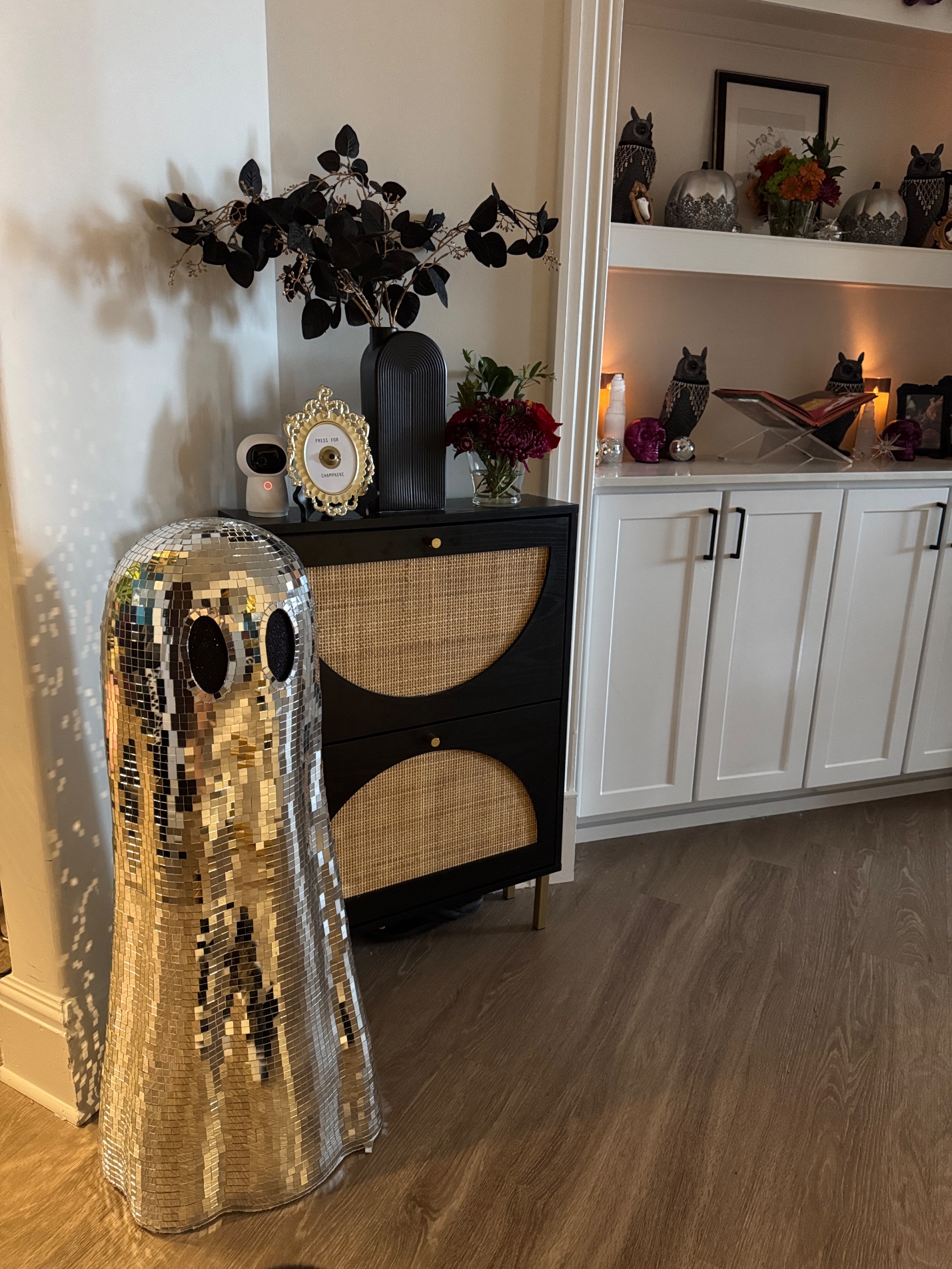 Disco ball ghost decoration beside a black cabinet with a Press for Champagne frame