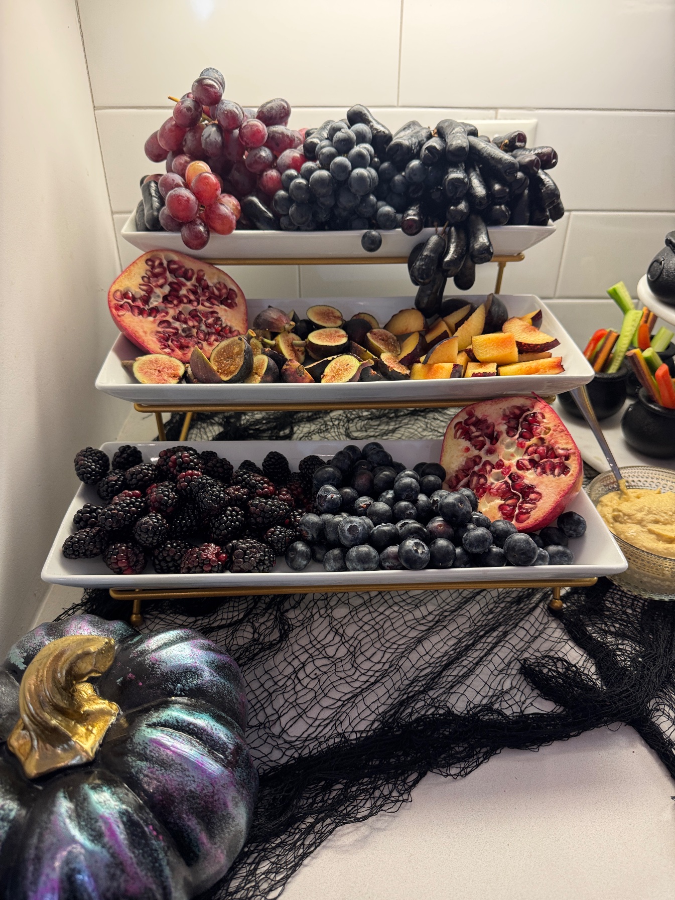 Three-tier fruit stand with moon grapes, black figs, plums, pomegranate, blackberries and blueberries