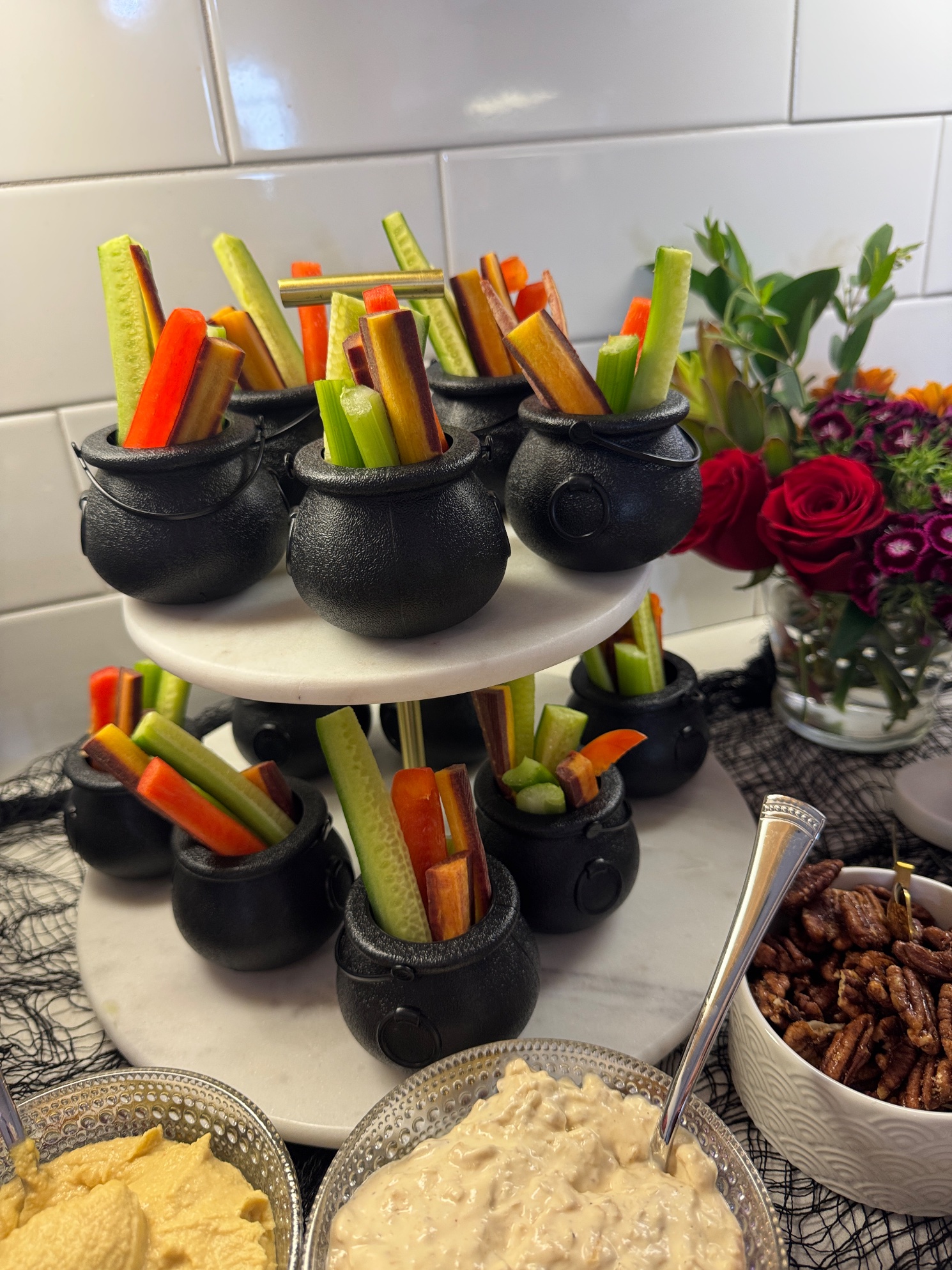 Veggie sticks in mini black cauldrons on a tiered marble stand with dips and pecans