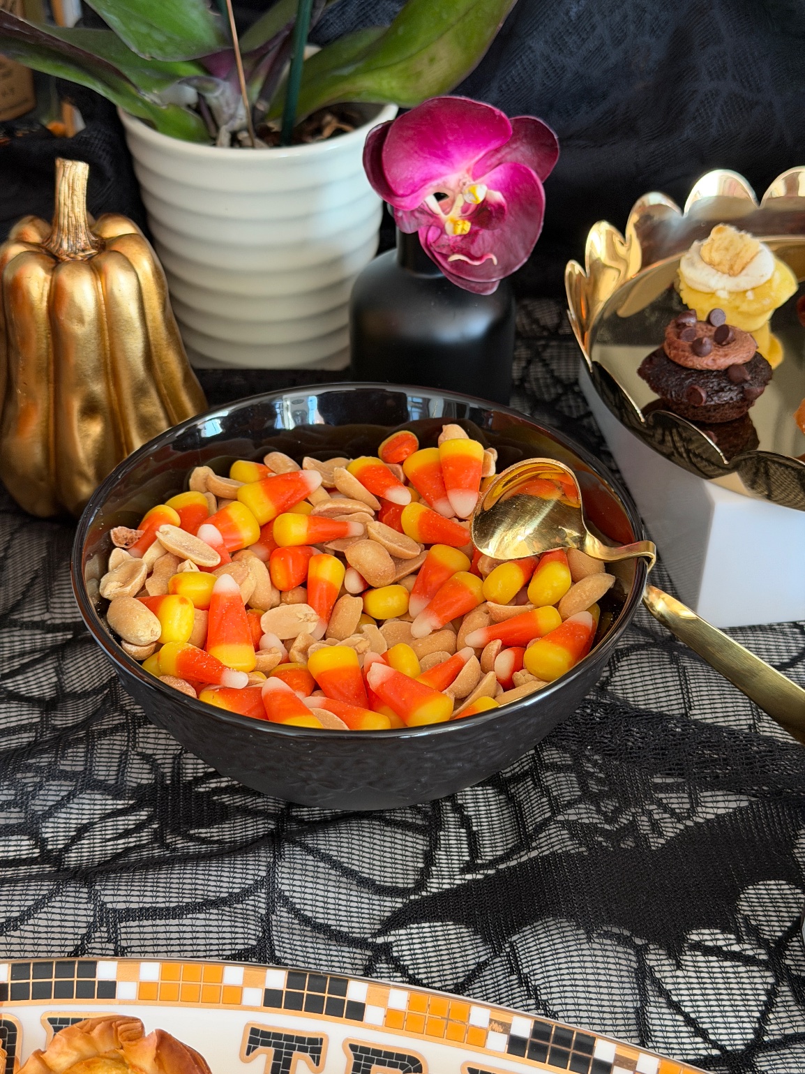 Black bowl of candy corn mixed with peanuts and a gold spoon