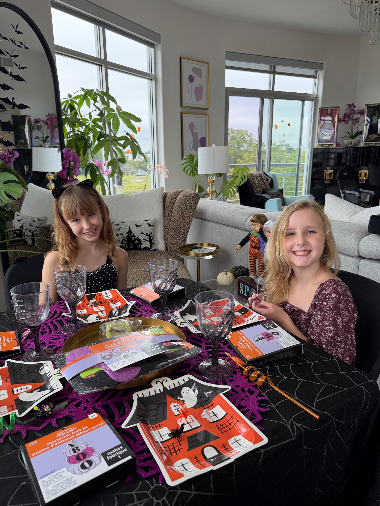 Lily and her friend at the kids' table with craft kits, dolls in Halloween outfits behind them