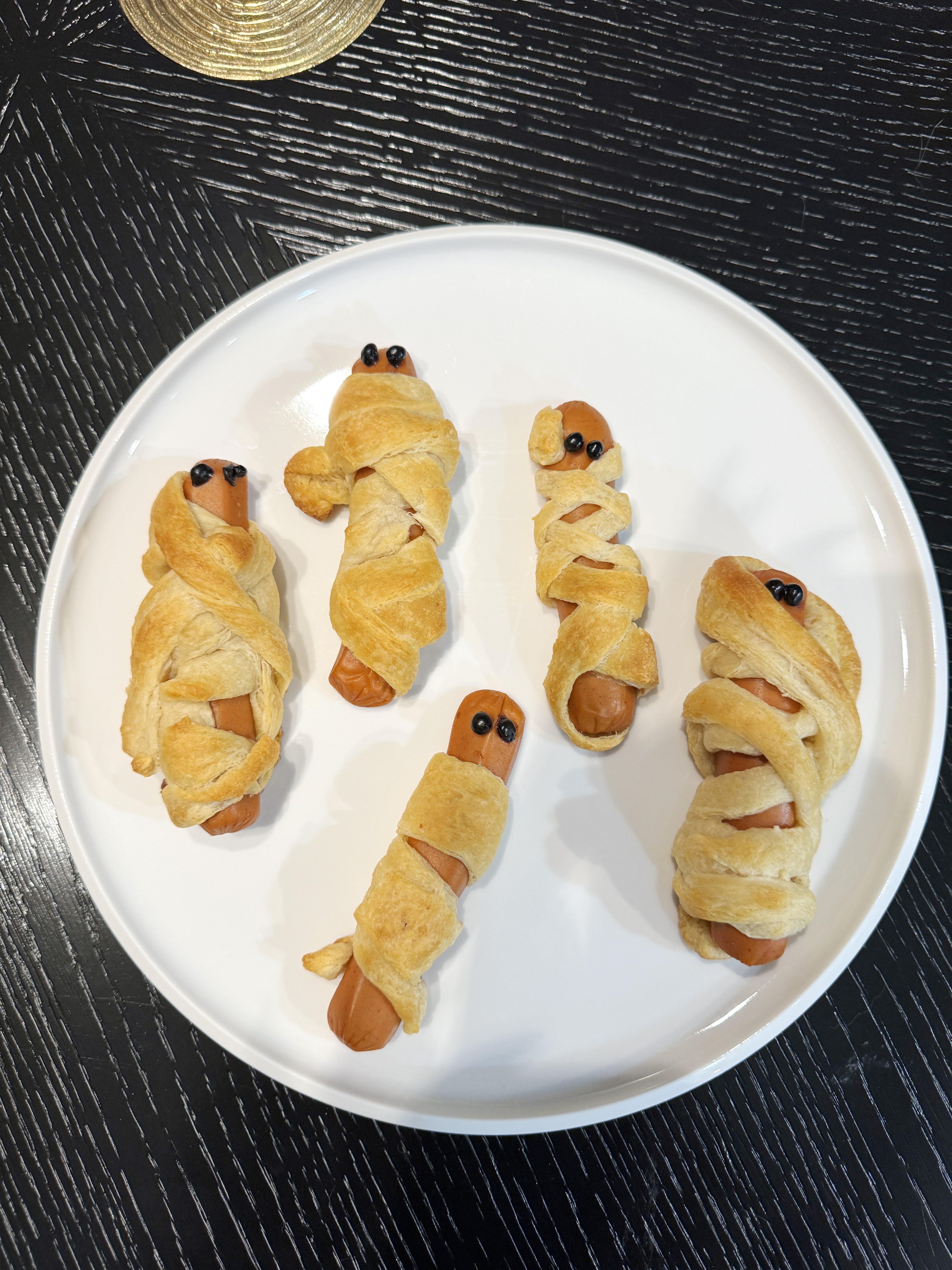 Mummy dogs on a white plate, hot dogs wrapped in crescent dough bandages with little dot eyes