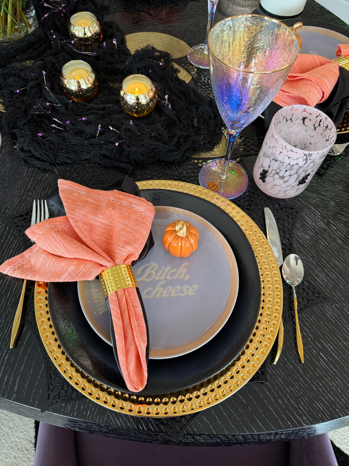 Galloween place setting with a gold beaded charger, black plate, lilac Bitch, cheese plate, mini pumpkin and coral napkin