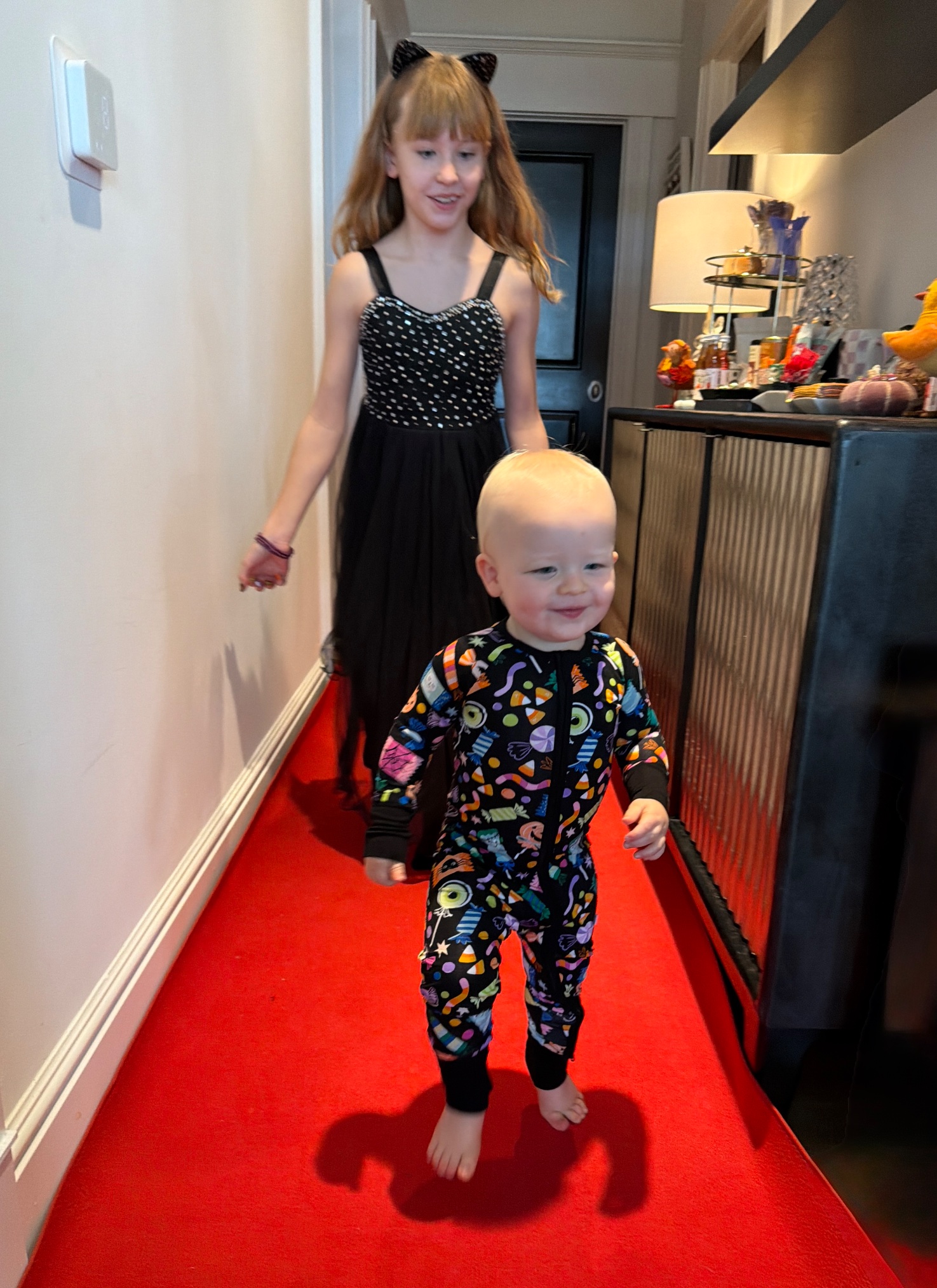Baby London toddling down the red carpet runner in his Halloween candy onesie, Lily behind him in her sparkle dress and cat ears